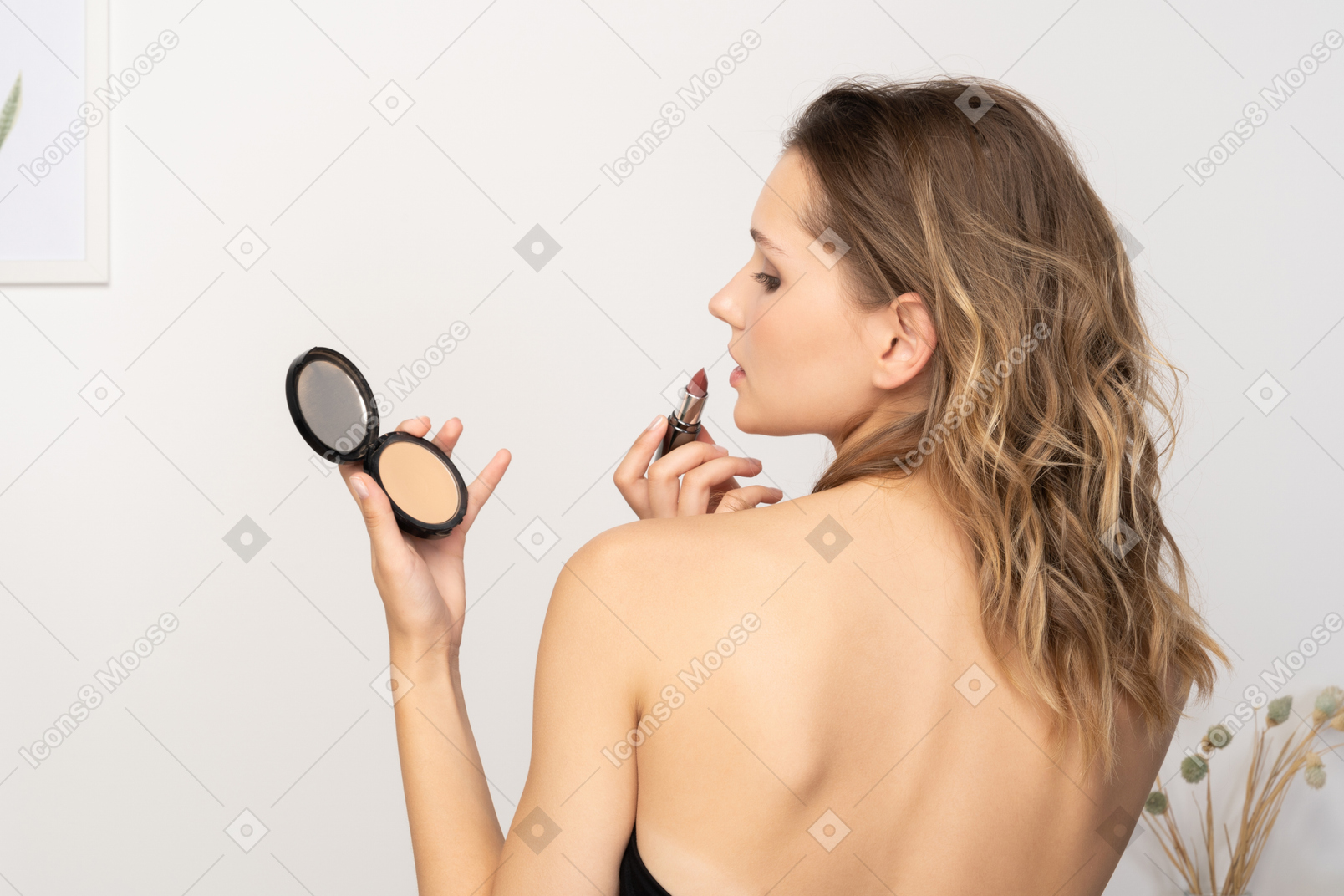 Dreiviertel-rückansicht einer sinnlichen jungen frau, die lippenstift aufträgt, während sie einen spiegel hält
