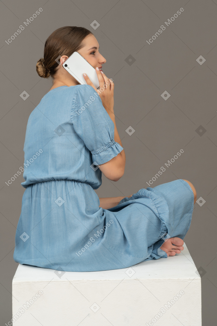 Vista laterale della giovane donna seduta sul cubo e parlando su smartphone