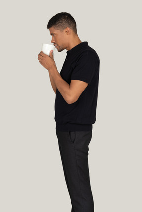 Standing in profile young man in black t-shirt and pants drinking coffee