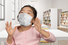 Fröhliches kleines mädchen mit gesichtsmaske, das in einer bibliothek steht