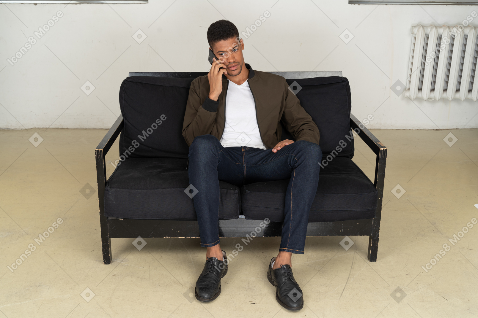 Vista frontale di un giovane perplesso seduto su un divano e parlando al telefono