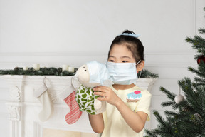 A little girl in face mask holding a puppet toy in face mask on christmas