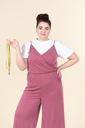 Jeune femme de taille plus dans une combinaison rose, posant avec un ruban à mesurer sur un fond jaune pastel