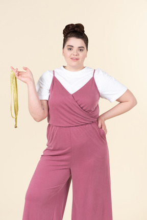 Jovem plus size em um macacão rosa, posando com uma fita métrica contra um fundo amarelo pastel