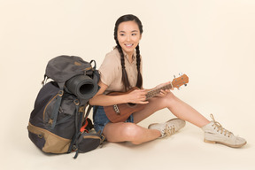 Lächelndes junges asiatisches mädchen, das nahe rucksack sitzt und die gitarre spielt