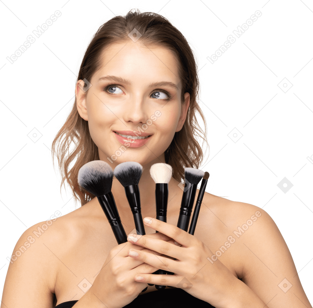 Front view of a smiling young woman holding make-up brushes and tilting head