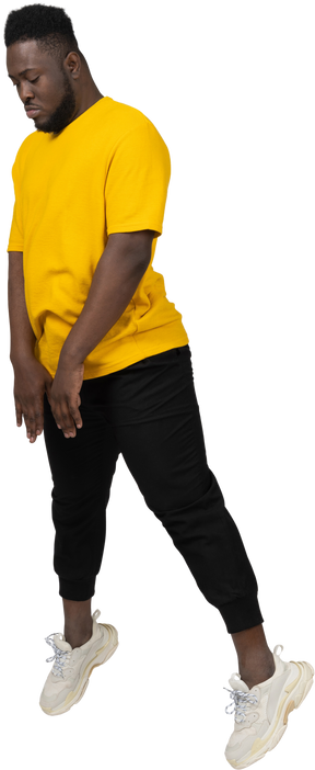 노란색 티셔츠를 입은 검은 피부가 아래를 내려다보고 있는 점프하는 젊은 남자의 3/4 보기