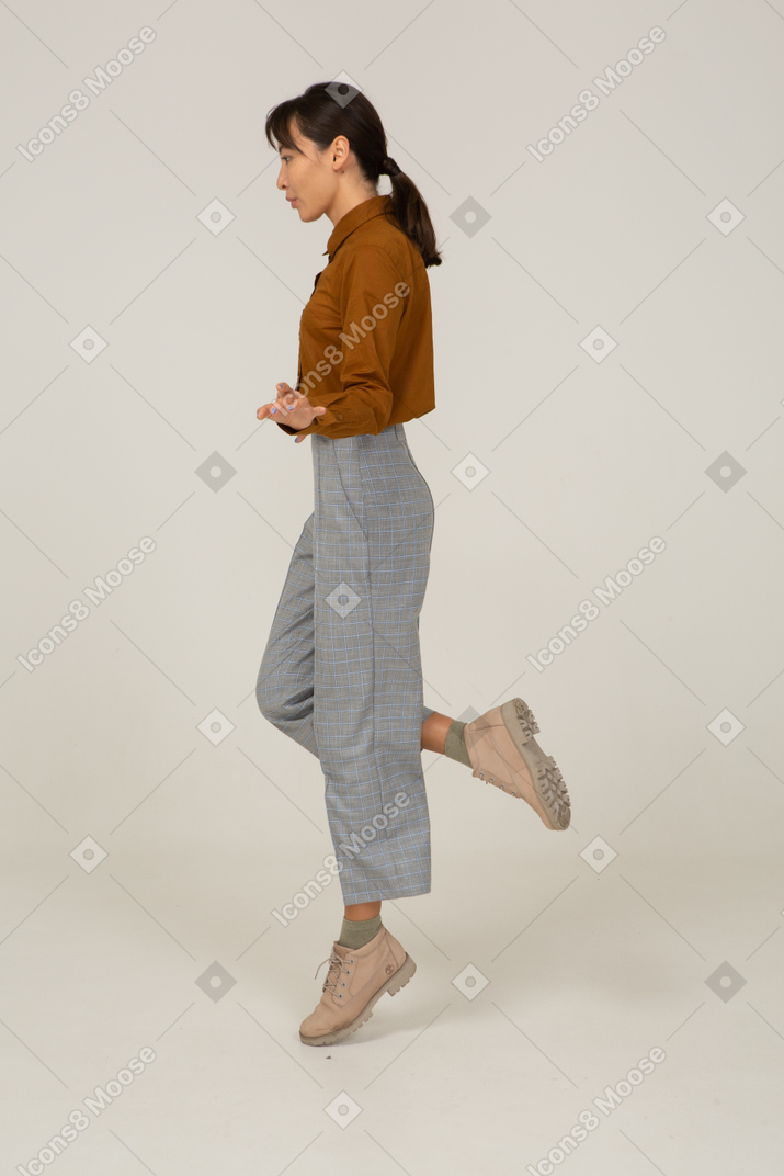 Vista lateral traseira de uma jovem fêmea asiática pulando de calça e blusa estendendo as mãos