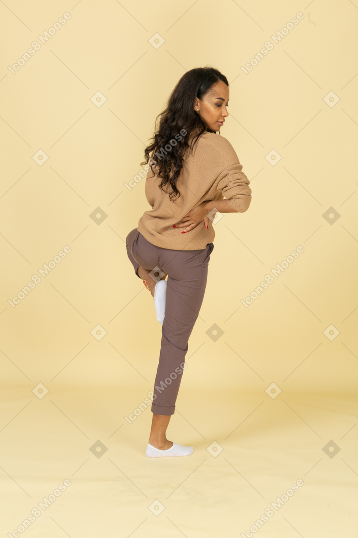 Three-quarter back view of a dark-skinned young female putting hand on hip while raising leg