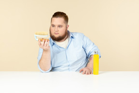 Househusband jovem com excesso de peso, sentado à mesa e olhando para a escova