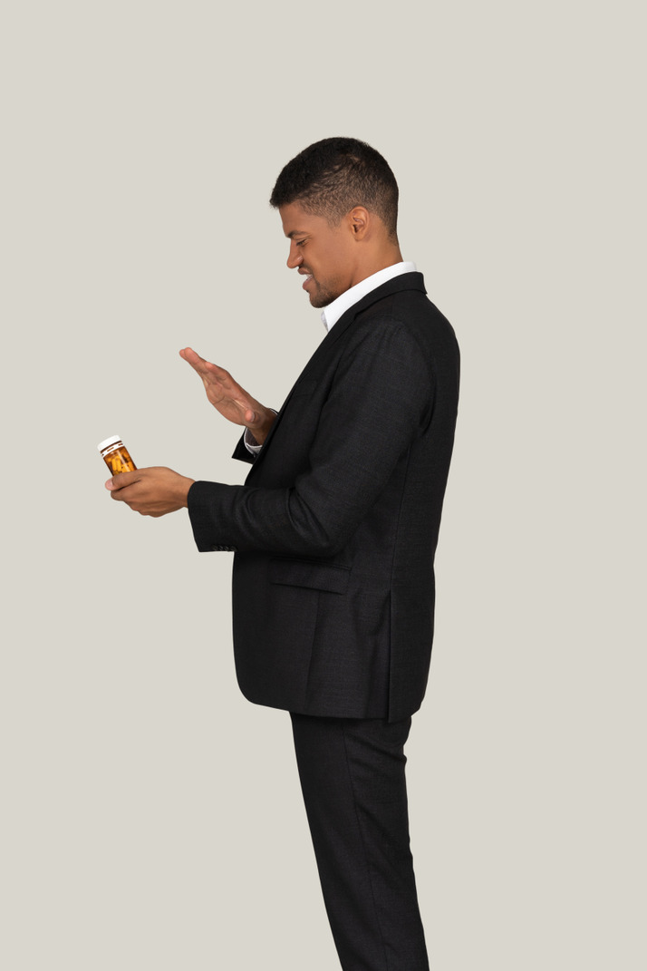Standing in profile young man in black suit looking at pills bottle he's holding with disgusted face