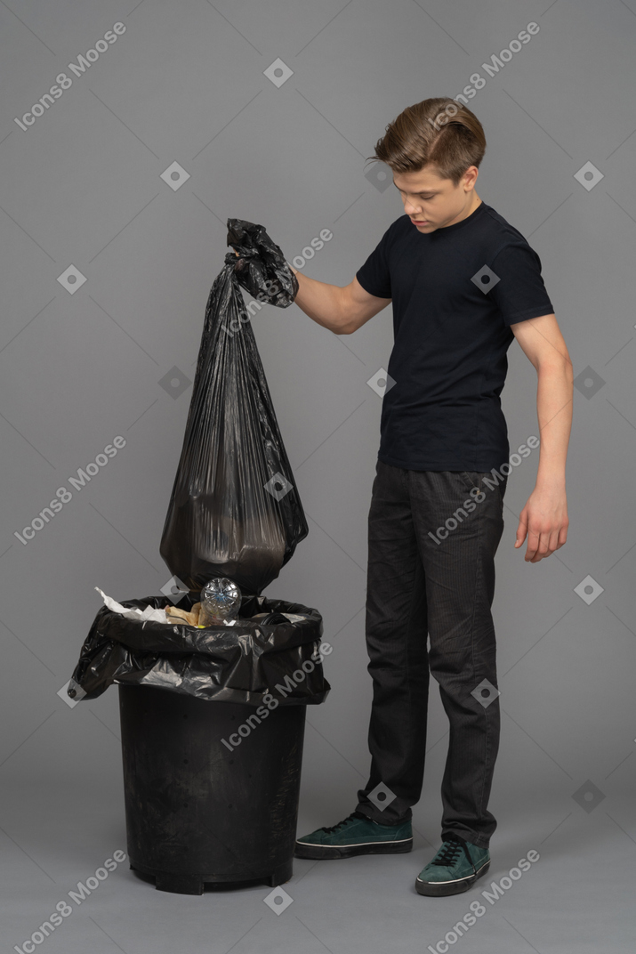 A young man holding a trash bag above a waste bin
