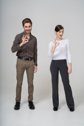 Vista frontal de um jovem casal com roupa de escritório mostrando sinal de ok