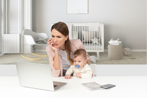 Eine schöne stirnrunzelnde frau, die durch den bürotisch mit einem baby sitzt und zum laptop schaut