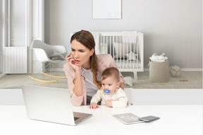 A beautiful frowned woman sitting by the office table with a baby boy and looking to laptop