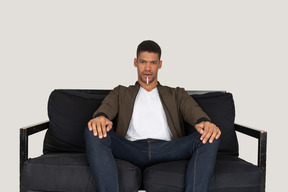 Front view of young man sitting on a sofa and holding cigarette in mouth