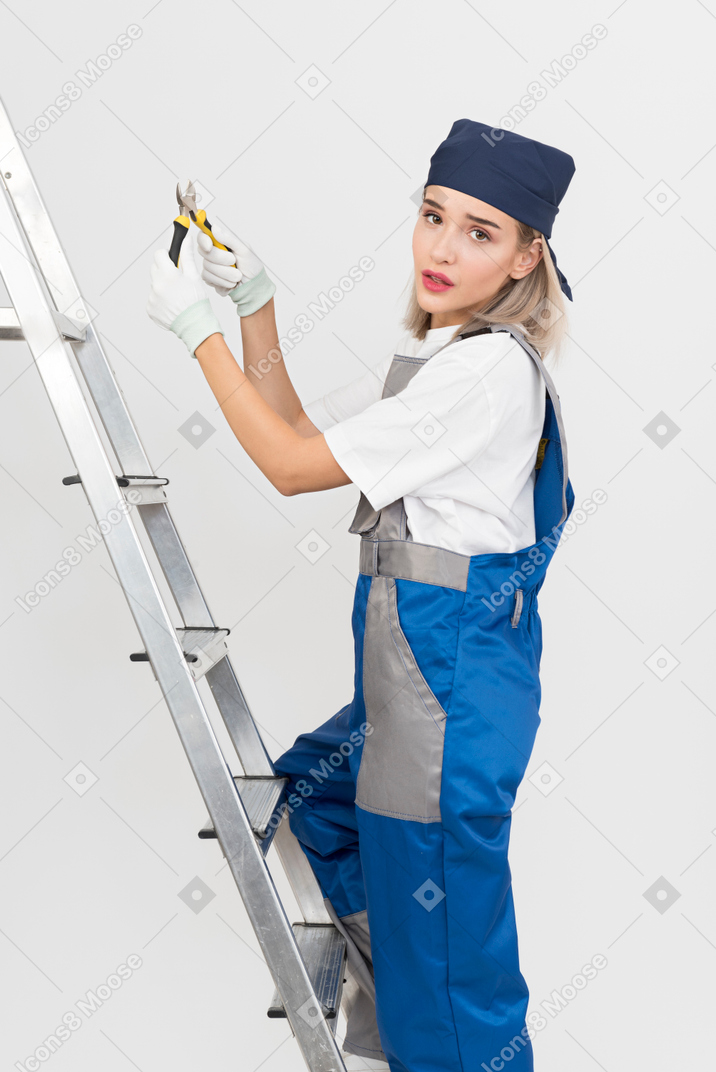 Trabajadora de pie en la escalera de tijera y sosteniendo alicates