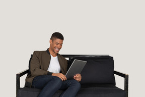 一个微笑的年轻人坐在沙发上看平板电脑的前视图
