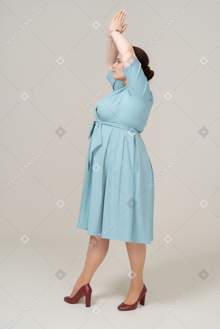머리 위로 손을 들고 서 있는 파란 드레스를 입은 여자의 측면 보기