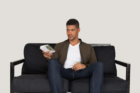 Front view of young man sitting on a sofa and passing pensil with notebook