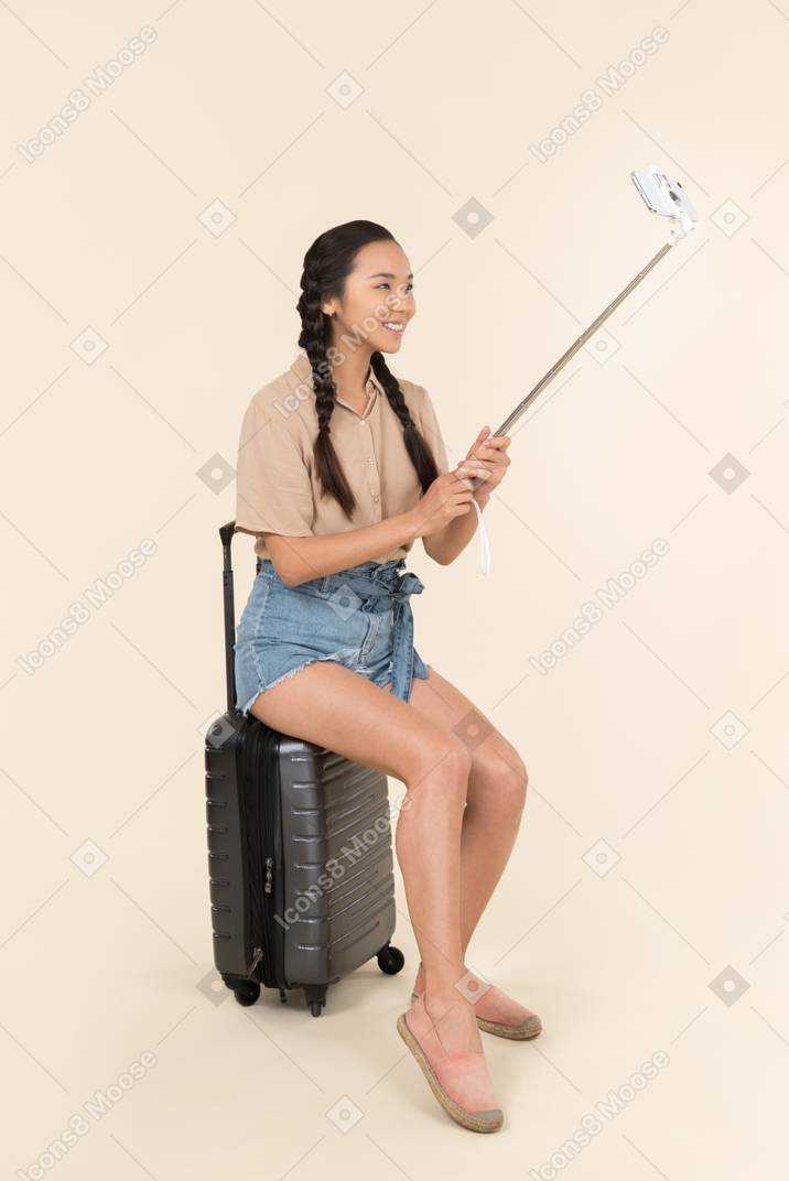 Young female traveller sitting on suitcase and holding selfie stick