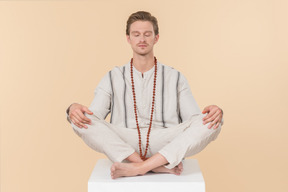 Young caucasian man with necklace sitting in lotus pose with his eyes closed