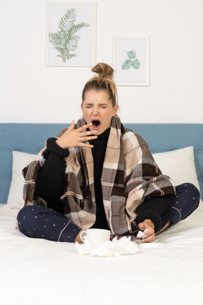 Vue de face d'un bâillement malade jeune femme en pyjama enveloppé dans une couverture vérifiée au lit