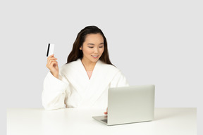 A woman in a bathrobe holding a credit card in front of a laptop