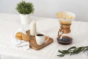 Chemex, grande tazza di caffè, biscotti e bottiglia di latte sul vassoio e pianta in vaso