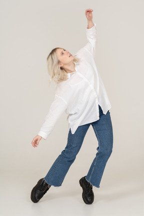 Front view of a blonde female in casual clothes dancing on her tiptoes and raising hand while looking up