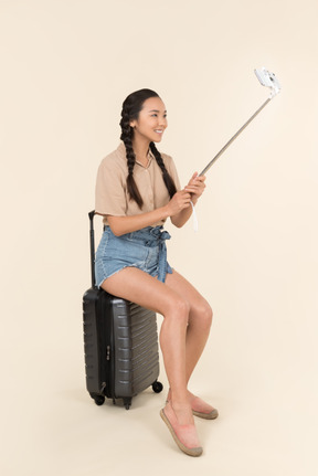 Young female traveller sitting on suitcase and holding selfie stick