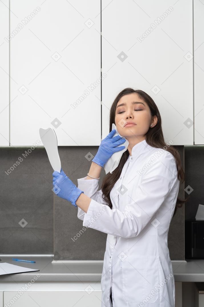 Vista di tre quarti di una dottoressa in possesso di uno specchio e asciugandosi il viso