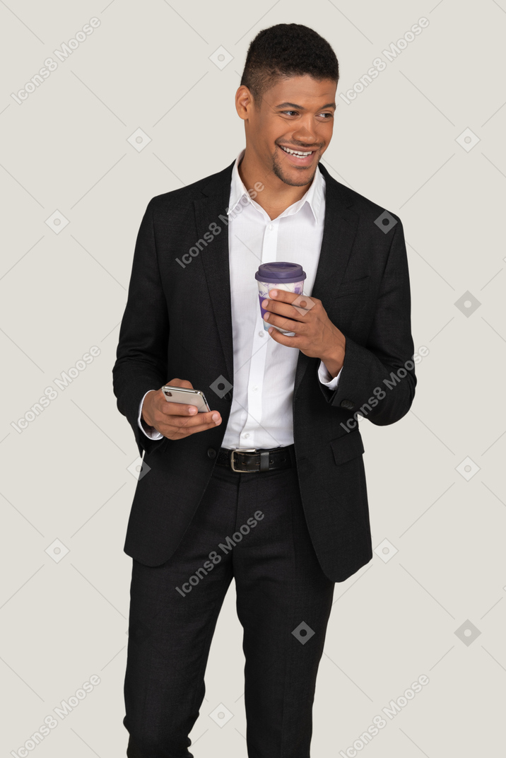 A handsome man dressed in the black suit and holding the cup of coffee and the phone