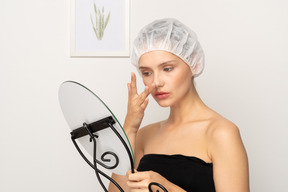 Mulher de touca cirúrgica segurando espelho e tocando seu nariz