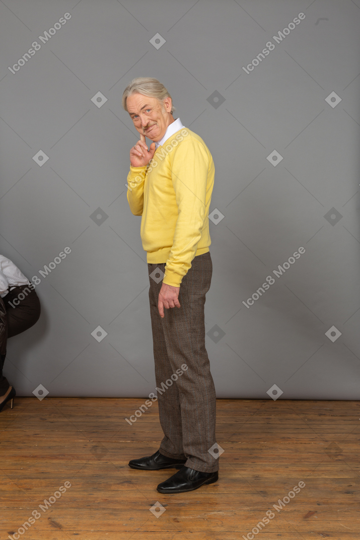 一个开朗的老人，在一件黄色的套衫，看着相机，摸脸的侧视图