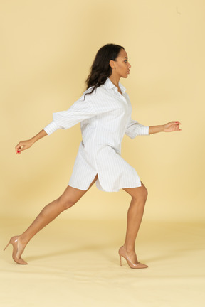 Vue latérale d'une jeune femme à la peau foncée en robe blanche faisant un grand pas