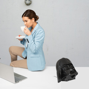 A woman sitting on a table with a laptop and a darth vader helmet