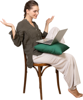 Vista de três quartos de uma jovem vestindo roupas de casa, sentada em uma cadeira com um laptop e levantando as mãos