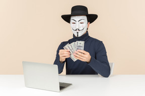 Hacker mit vendetta-maske am tisch sitzen und geld zählen