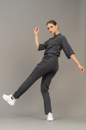 Side view of a young woman in a jumpsuit outstretching her arm & raising leg