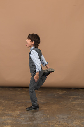 Vista lateral de un niño con traje gris posando en una pierna