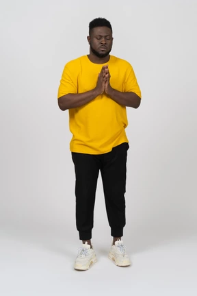 Front view of a praying young dark-skinned man in yellow t-shirt holding hands together