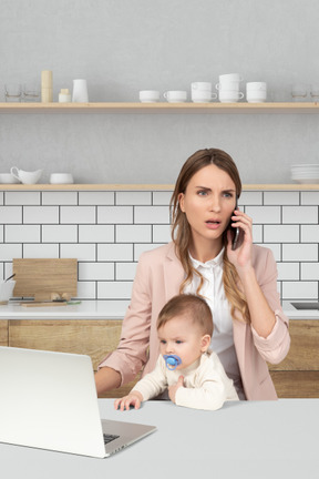 A woman with baby sitting at a table with laptop and talking on a phone