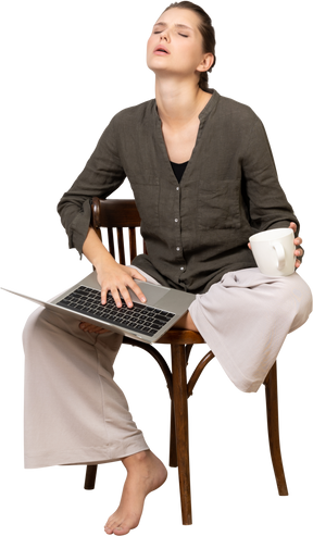 Front view of a tired young woman wearing home clothes sitting on a chair with a laptop & coffee cup