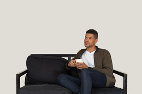 Front view of a young dreaming man sitting on a sofa with a cup of coffee