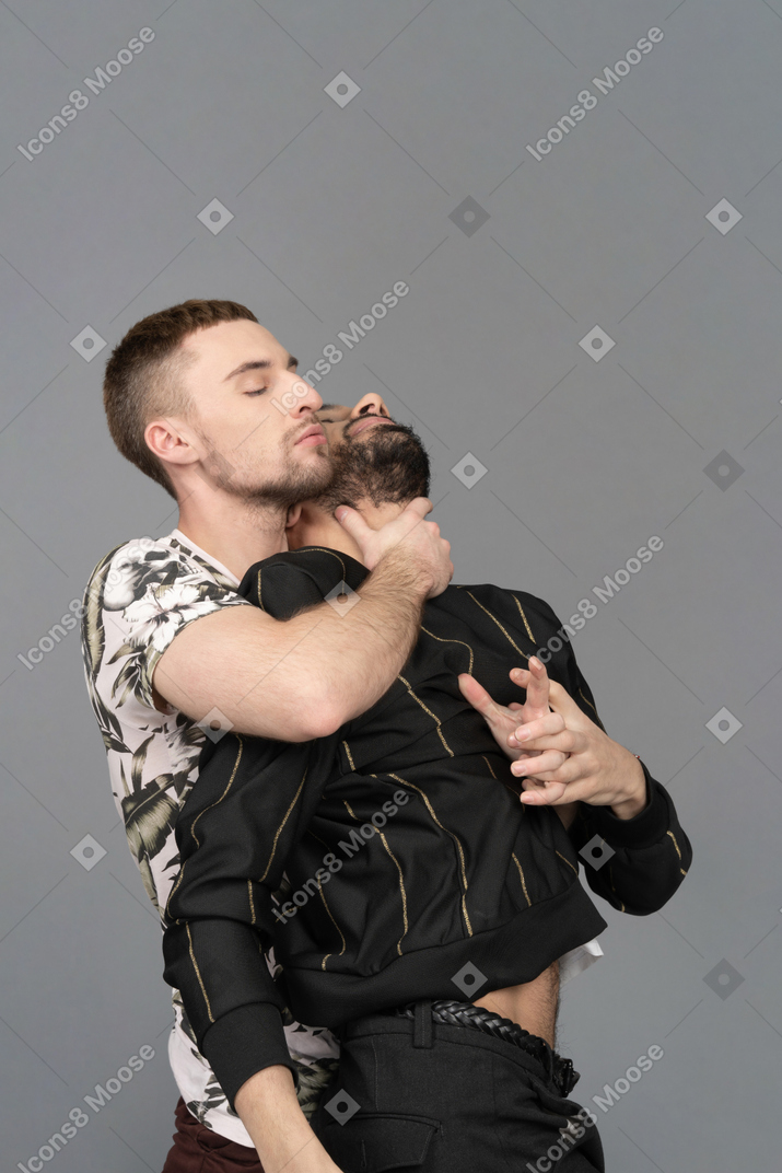年轻的白人男子从背后性感地拥抱他的伴侣，一只手放在他的脖子上