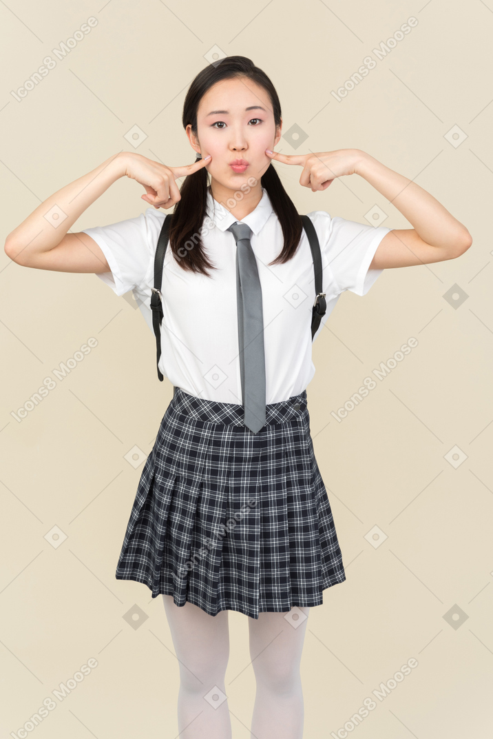 Asian school girl making duck face and pointing at her cheeks with fingers