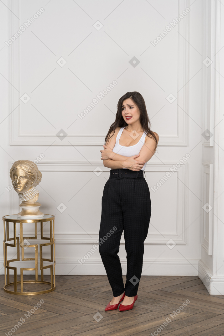 Гримасничающая молодая женщина в полный рост, скрещивающая руки, стоя возле золотой греческой скульптуры