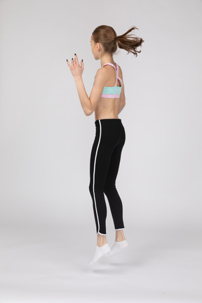 Vista posterior de tres cuartos de una jovencita en ropa deportiva levantando la mano y saltando