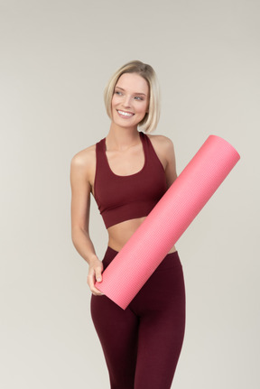 Улыбающаяся молодая женщина в спортивной одежде держит коврик для йоги